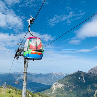 Bunte Gondel mit Ramsi-Maskottchenmotiv schwebt über grüne Almwiesen und Berge im Sommer.