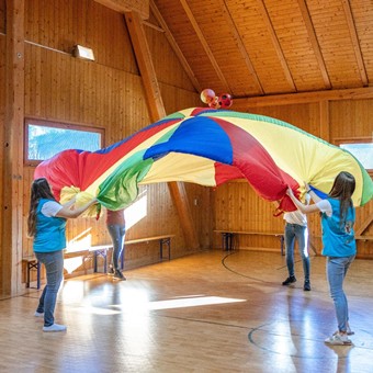 Four women swing a colorful swing cloth with three balls in the bright gymnasium of Hotel Ramsi.