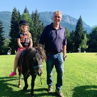 Mädchen reitet auf einem braunen Pony über eine grüne Wiese, begleitet von einem freundichen Ramsi-Mitarbeiter vor vielen Bäumen und Bergkulisse.