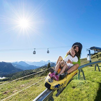 Mom and daughter summer tobogganing in front of gondolas hovering over a mountain backdrop in bright sunshine.
