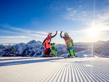 Mutter und Vater, geben sich ein High five auf einer Skipiste während ihre drei Kinder daneben stehen und zusehen.