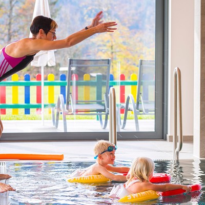 Two children in a pool, swimming with the help of buoyancy aids and swimming boards, try to fetch the balls that a woman throws from the edge of the pool.