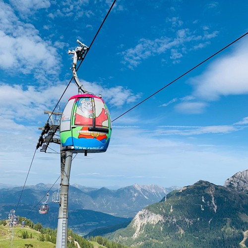 Bunte Gondel mit Ramsi-Maskottchenmotiv schwebt über grüne Almwiesen und Berge im Sommer.