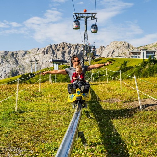 Vater und Sohn genießen die rasante Fahrt mit der Sommerrodelbahn inmitten der Berge unter Gondeln.