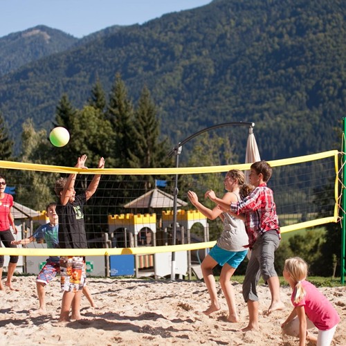 Kinder und Teenager spielen gemeinsam Beachvolleyball vor einer Bergkulisse und dem Abenteuerspielplatz des Hotels Ramsi.