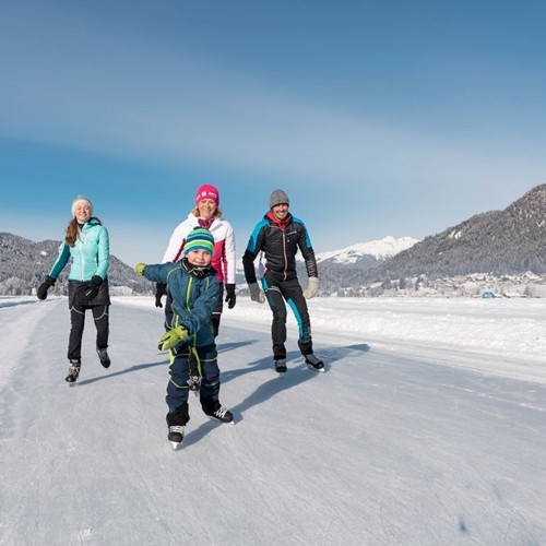 Familie beim Eislaufen vor einer Bergkulisse und unter strahlend blauem Himmel.
