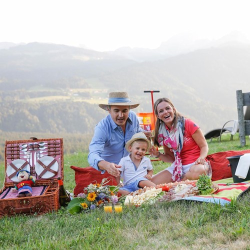 Family and Ramsi mascot dolls enjoying a picnic on the alpine meadow with panoramic views and regional delicacies.