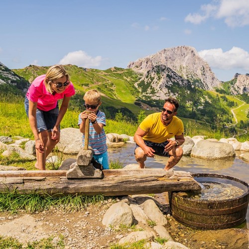 Familie beim Spielen mit einer Wasserspielstation inmitten der Alpenlandschaft.