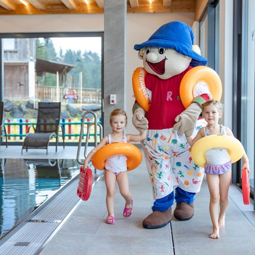 Two girls with Ramsi mascot, swimming boards and swimming rings laugh as they walk hand in hand along the edge of the indoor pool at Hotel Ramsi.